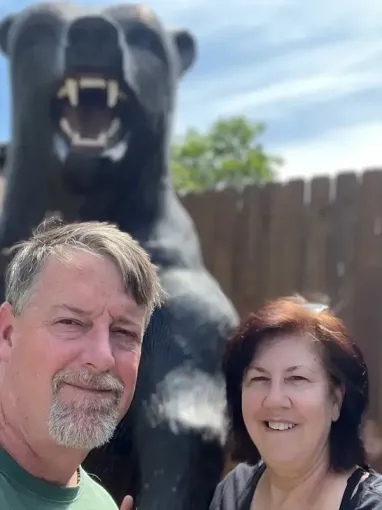 Smiling couple posing in front of large roaring bear statue outdoors.
