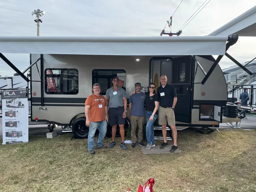 Group of people posing in front of PLA travel trailer at RV show.
