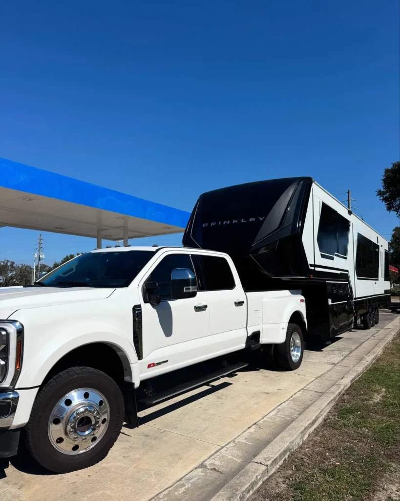 White Ford dually truck towing large Brinkley fifth-wheel RV near gas station.